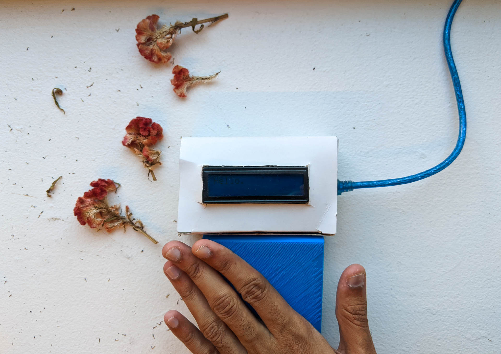 image of the first prototype, which is a white card paper box with a narrow blue LCD embedded within it. It has a 3D printed PLA square shaped panel, which contains the antenna connected to the box. It is currently deactivated, with a hand close to it